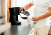 Person pouring coffee from carafe into mug beside drip coffee maker
