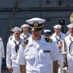 87 Cruise Crew Members Removed naval officer in white uniform stands before a military band