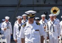 87 Cruise Crew Members Removed naval officer in white uniform stands before a military band